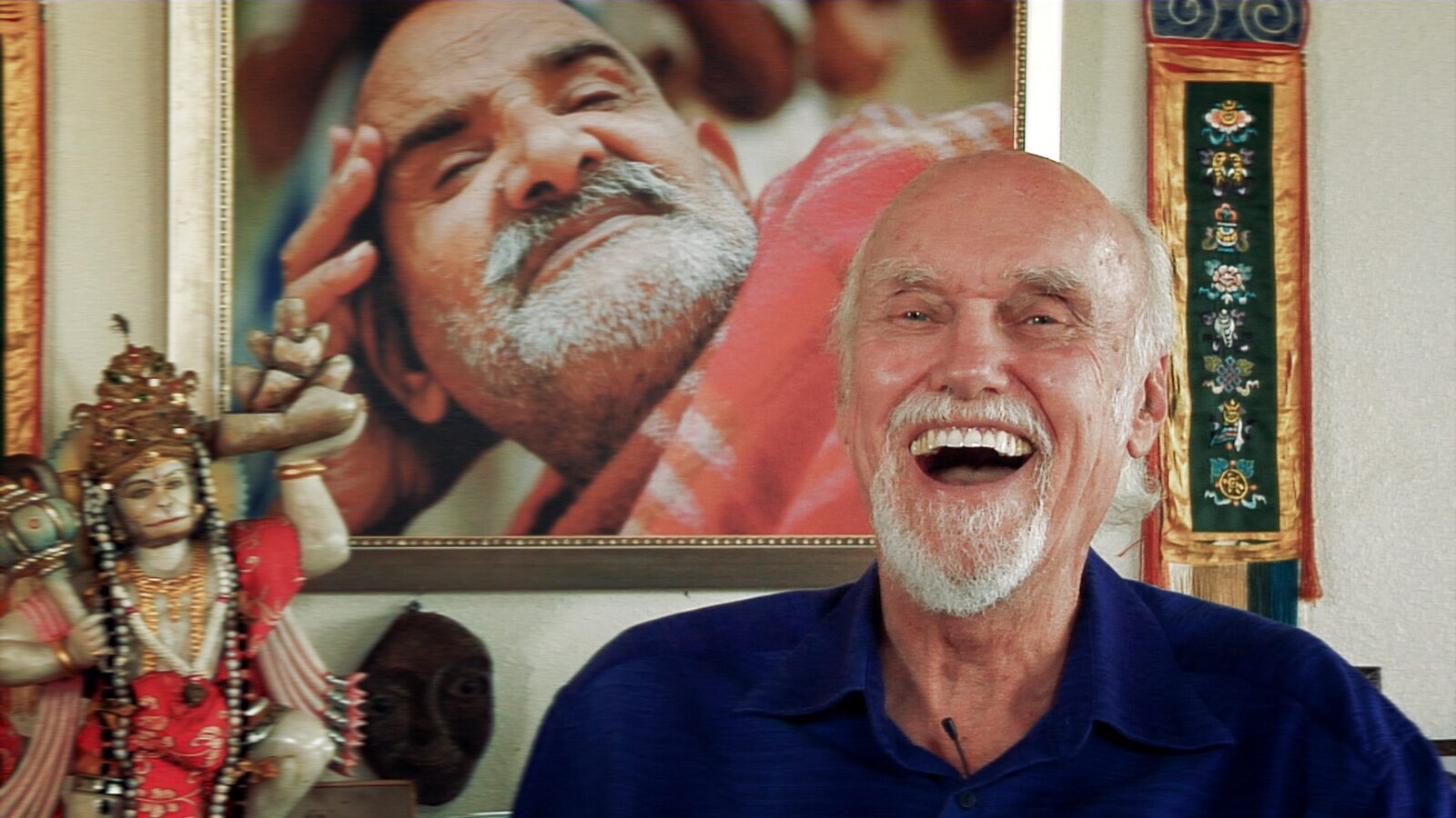An elderly man laughing joyfully in front of a large smiling portrait.